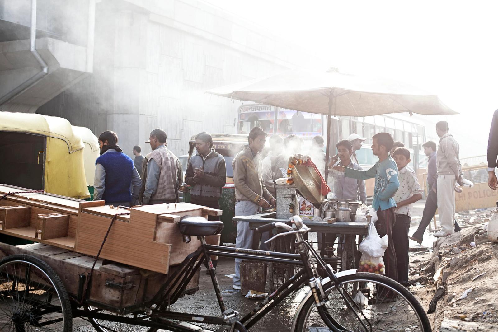 A bustling street scene in India, with a food cart and a crowd of people in the background, and a yellow auto-rickshaw and a black bicycle in the foreground.
