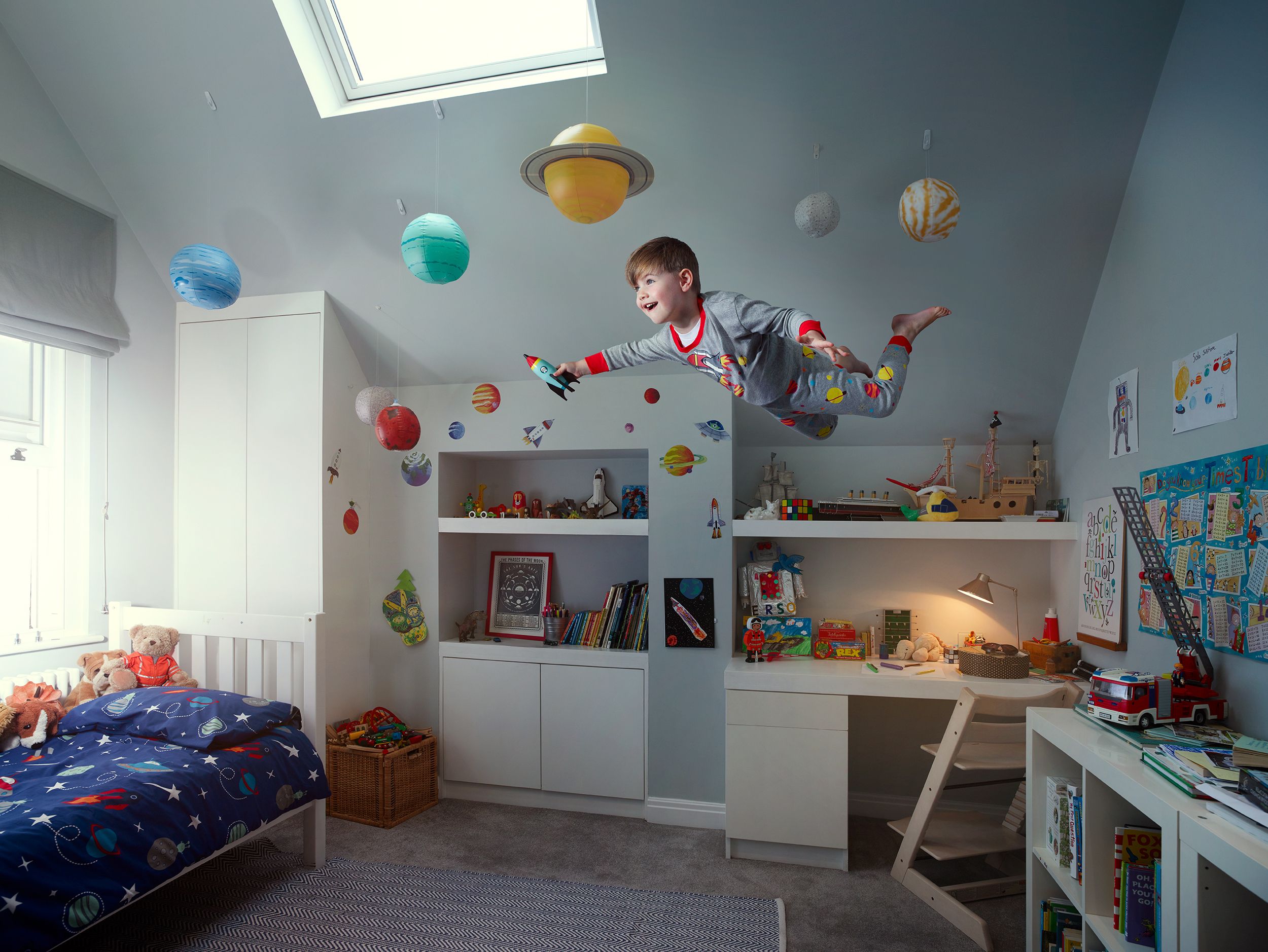 A young boy in a grey and red superhero costume is flying through the air in a bedroom filled with toys and books, surrounded by floating planets and stars.