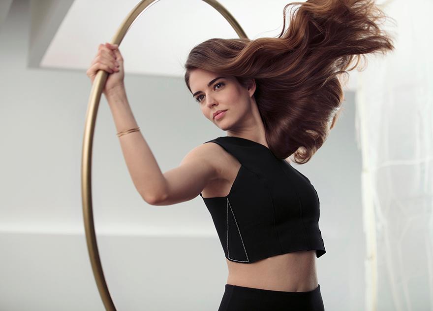 A woman in a black sports bra and leggings, holding a gold hula hoop and posing in a dynamic, athletic pose.