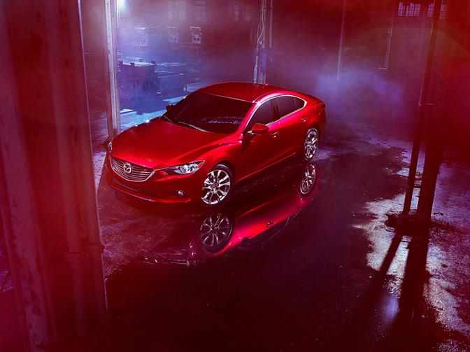 A red Mazda car is parked in a dimly lit, industrial-style garage with a backdrop of metal pipes and machinery.