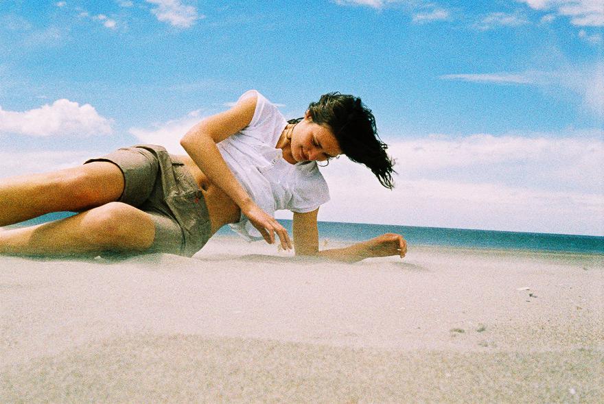 A woman is lying on a beach, wearing a white shirt and brown shorts, with her hair blowing in the wind.