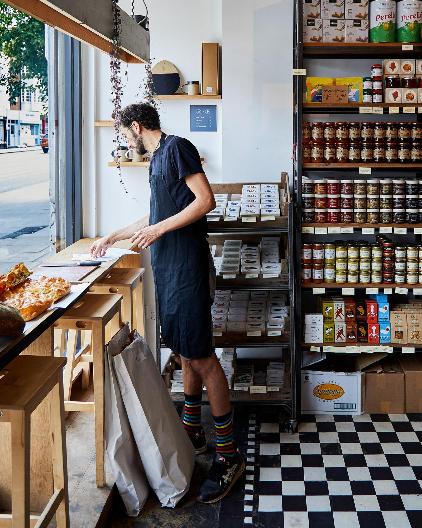A man in a black t-shirt and overalls is standing in a store, surrounded by shelves of food and products, possibly a food or grocery store.