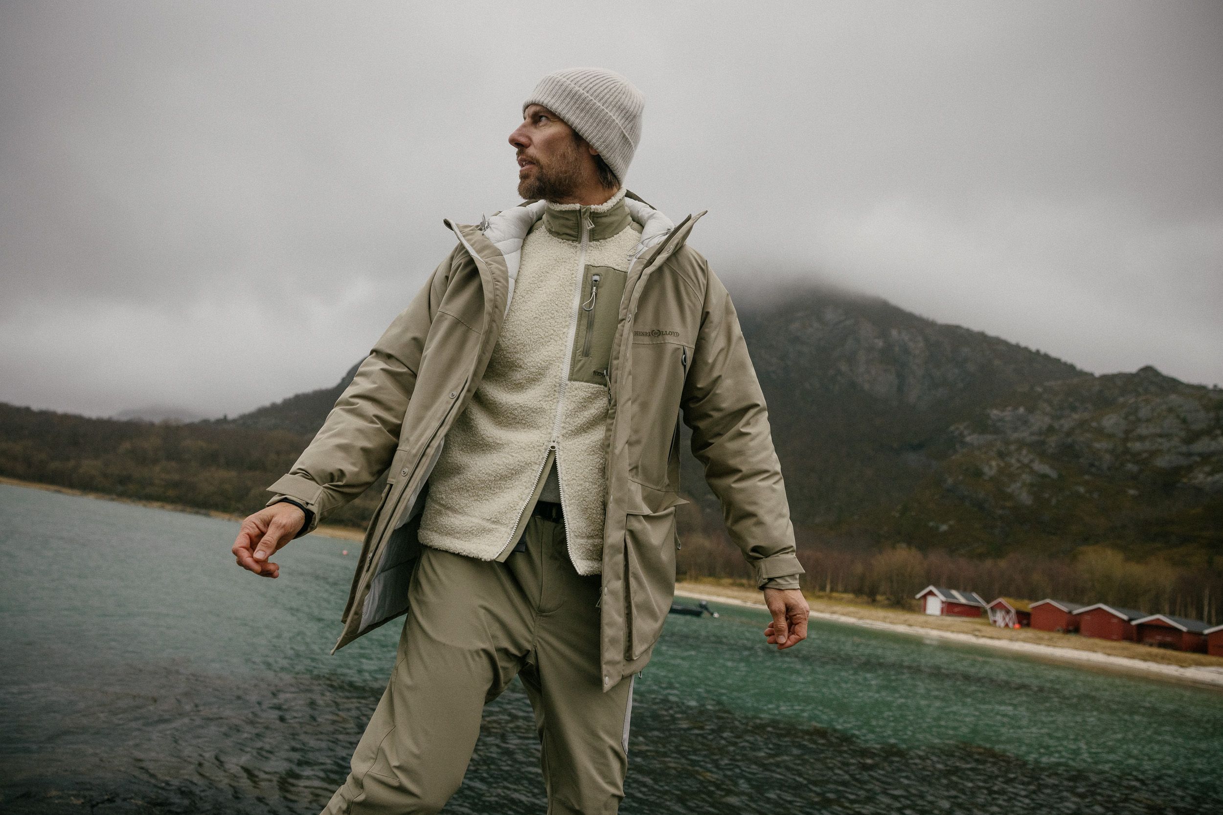A man in a white beanie, beige parka, and khaki pants stands on a rocky shoreline, gazing out at a misty mountain range.