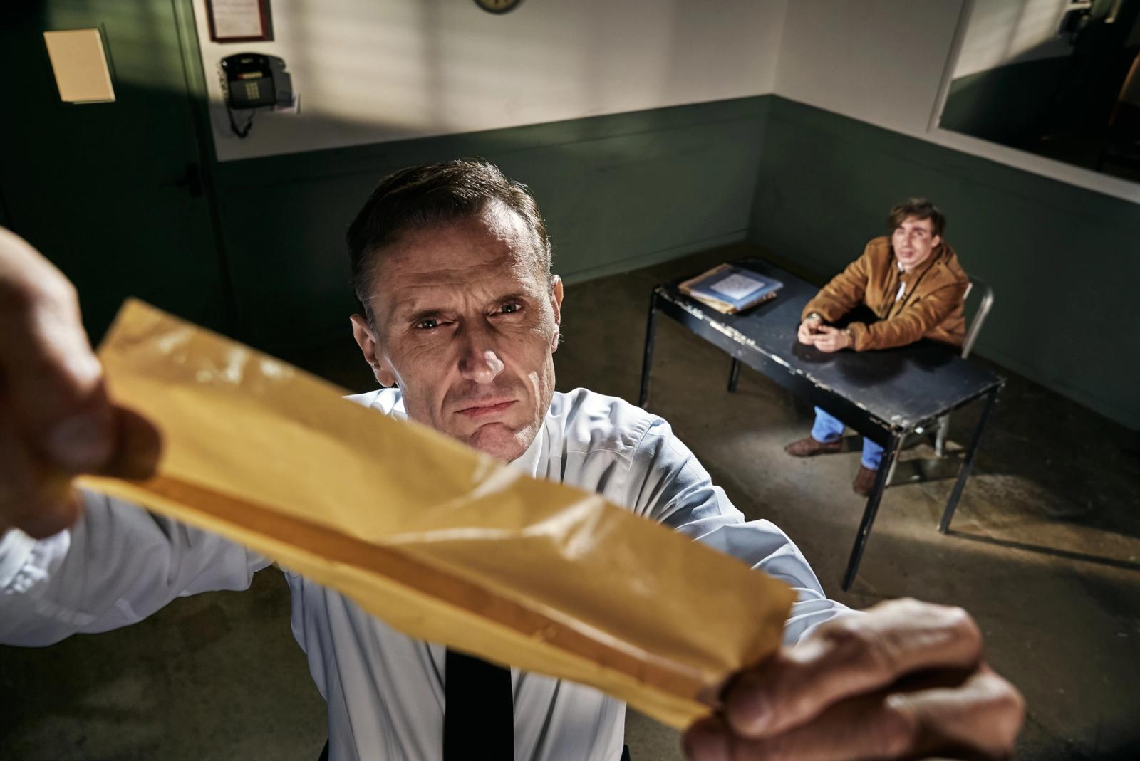 A man in a white shirt and black tie holding up a yellow envelope in a dimly lit room with another man sitting at a desk in the background.