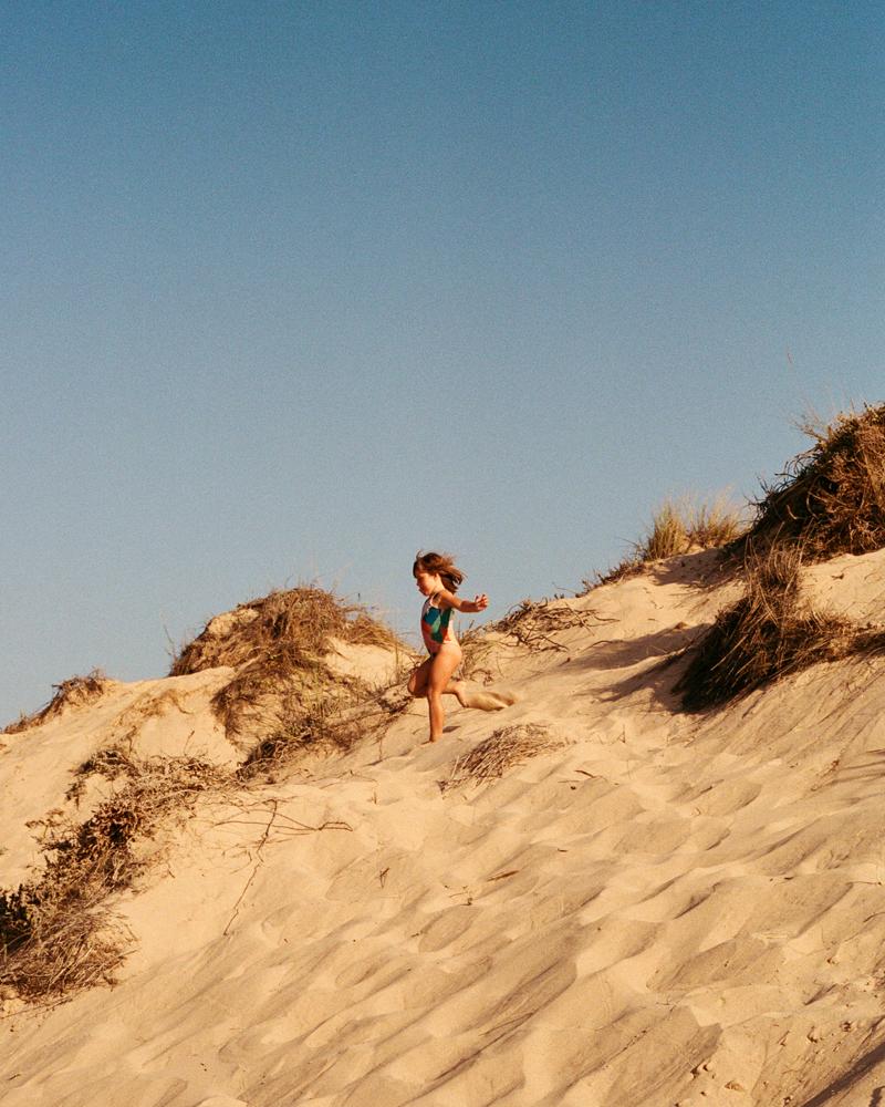 A woman in a swimsuit is running down a sandy dune, with the wind blowing through her hair.