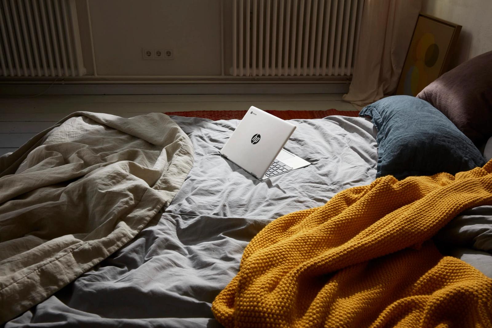 A dimly lit bedroom scene, with a laptop on a bed, creating an intimate and personal atmosphere. The laptop is placed on a bed, with a yellow blanket and a few pillows,.