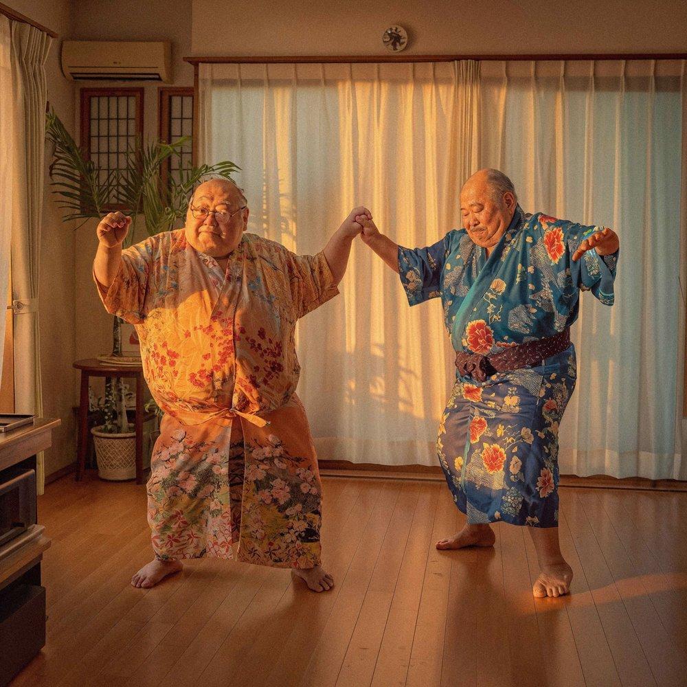 Two elderly men in traditional Japanese kimonos, holding hands and dancing in a living room with a wooden floor and a plant in the background.
