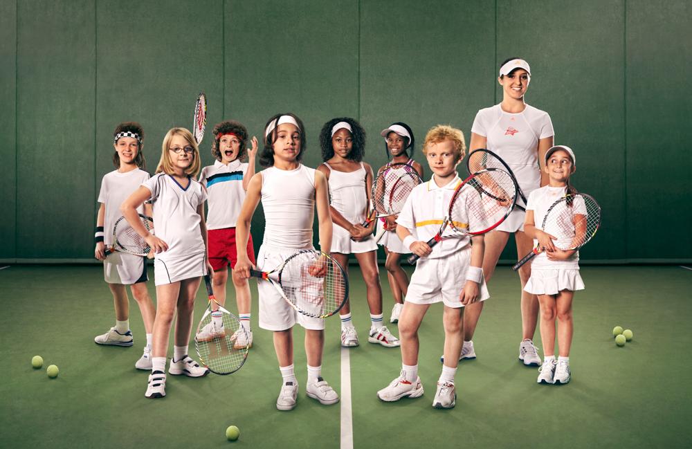 A group of children and a woman in tennis outfits, holding tennis rackets, standing on a tennis court with tennis balls scattered around them.