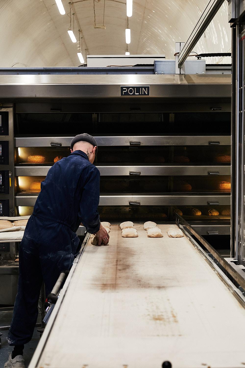 A man in a blue denim jumpsuit and a black beanie is working on a large industrial oven, placing bread rolls on a conveyor belt.