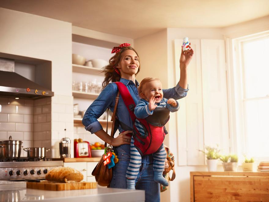 A woman wearing a denim shirt and jeans, holding a baby in a red baby carrier, standing in a kitchen with a stove and a counter with food and appliances.