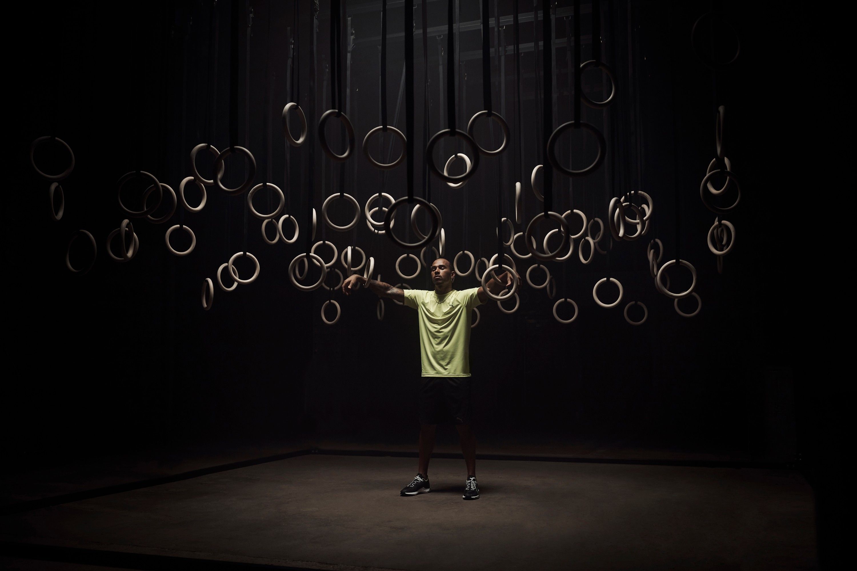 A man in a yellow t-shirt and black shorts stands with arms outstretched in front of a wall of rings.