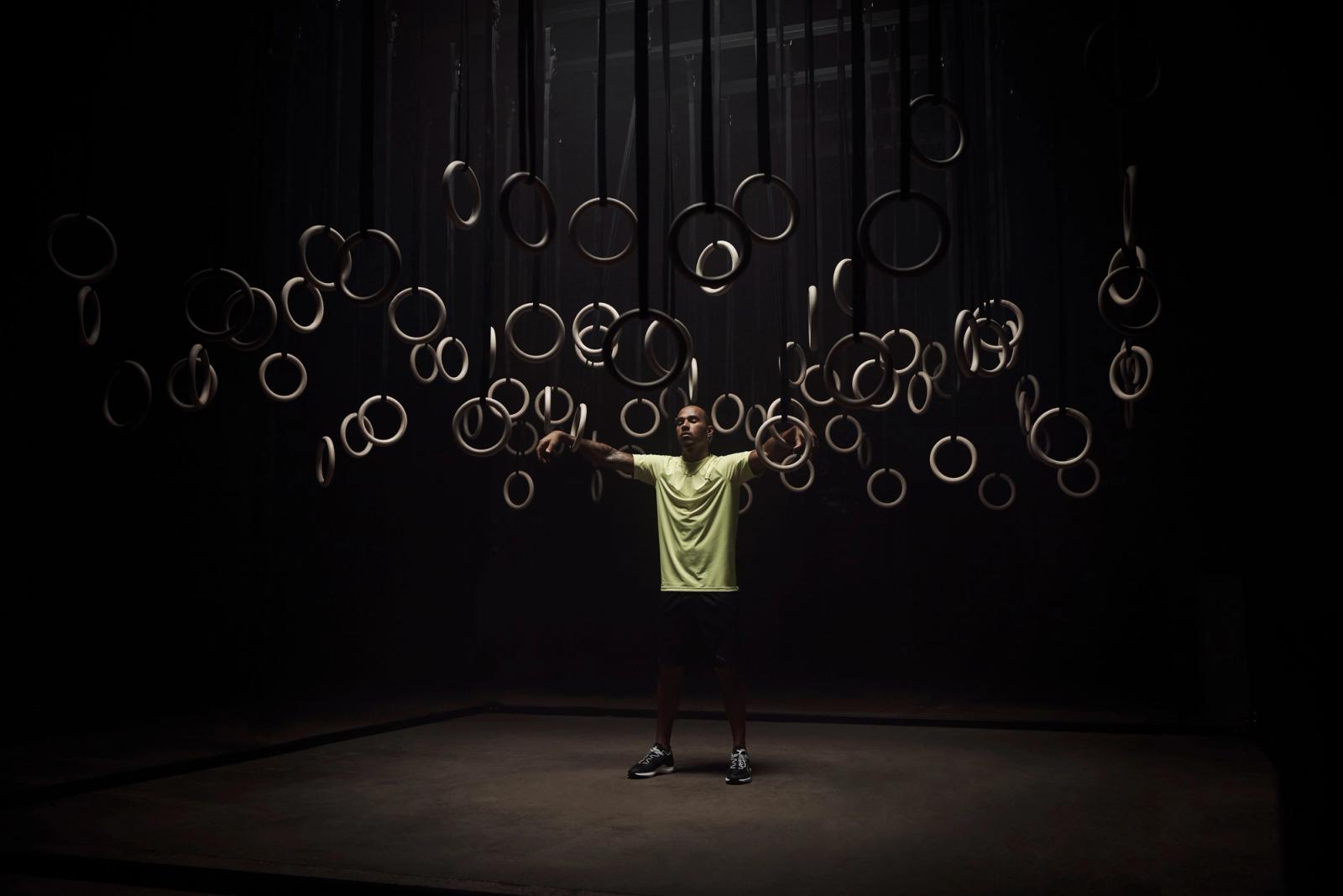 A man in a yellow t-shirt and black shorts stands with arms outstretched in front of a wall of rings.