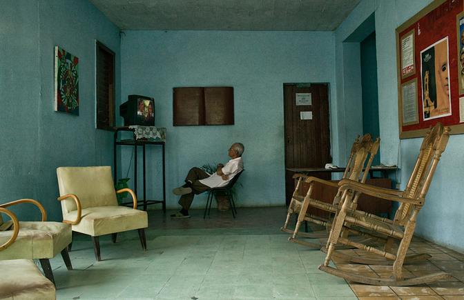 An elderly man sits in a waiting room, surrounded by worn, old-fashioned chairs and a small TV on a table in the corner.