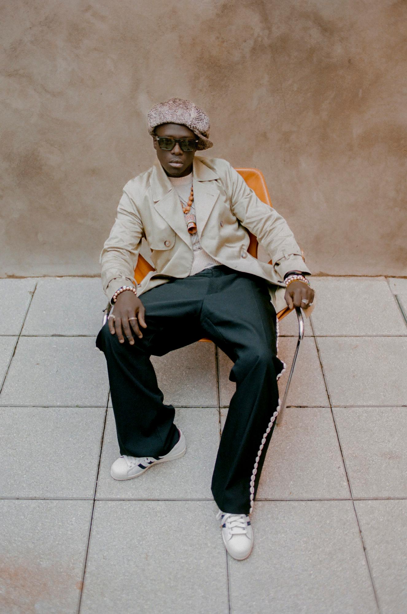 A man sitting on a chair, wearing a beige jacket, black pants, and a hat, with a chain around his neck and a watch on his wrist, in a modern, urban setting.
