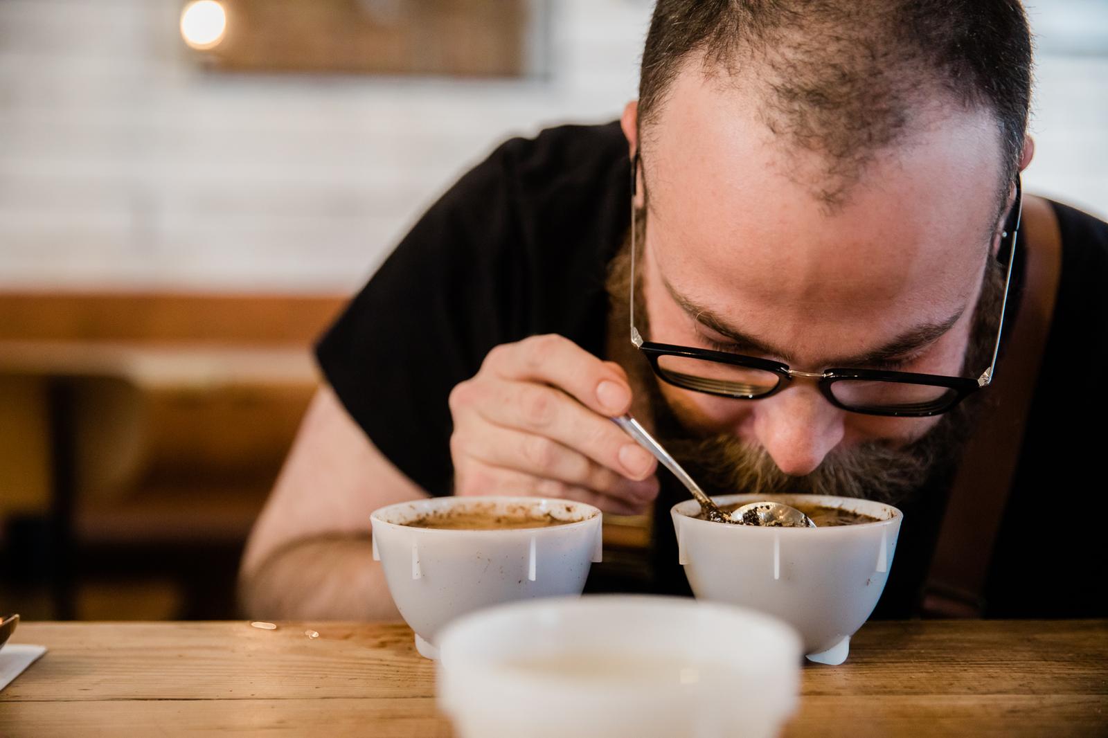 A man tastes coffee from a spoon in a cup.