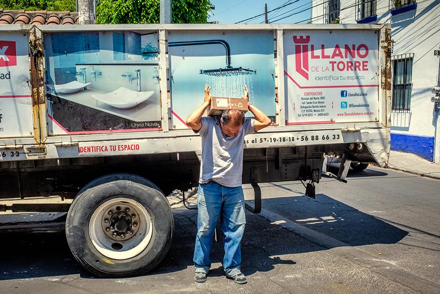 A man in a grey t-shirt and blue jeans is standing in front of a large white truck with a bathroom-themed advertisement on the side, holding a cardboard box on his head.