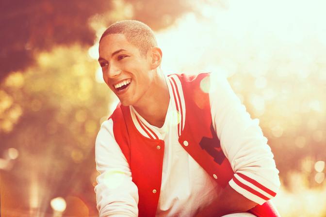 A young man with a big smile is wearing a red and white striped rugby jersey, a white baseball cap, and is holding a rugby ball in a park with trees and a green grassy field in the background.