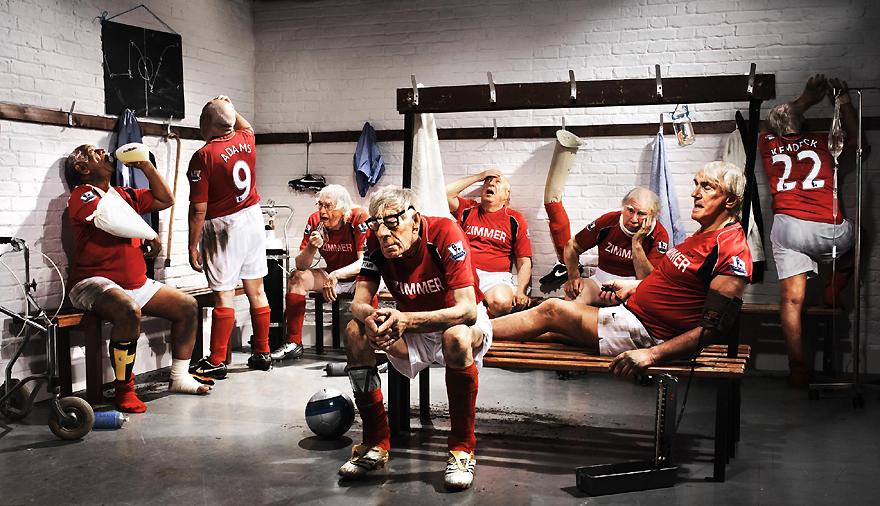 A group of older men sit on a bench in a locker room, some drinking from bottles, others using their phones.