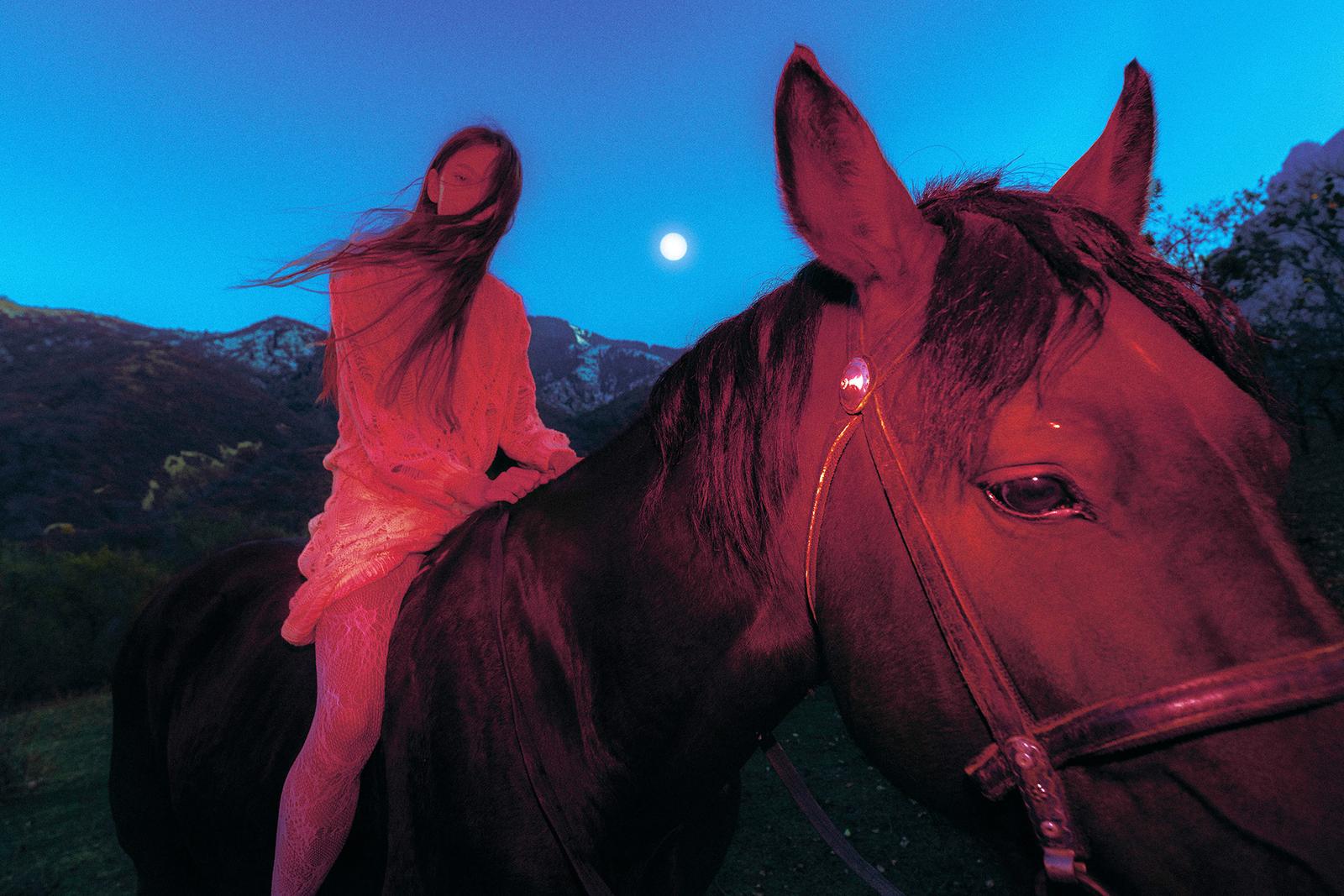 A woman in a white dress riding a horse in a mountainous landscape at sunset.