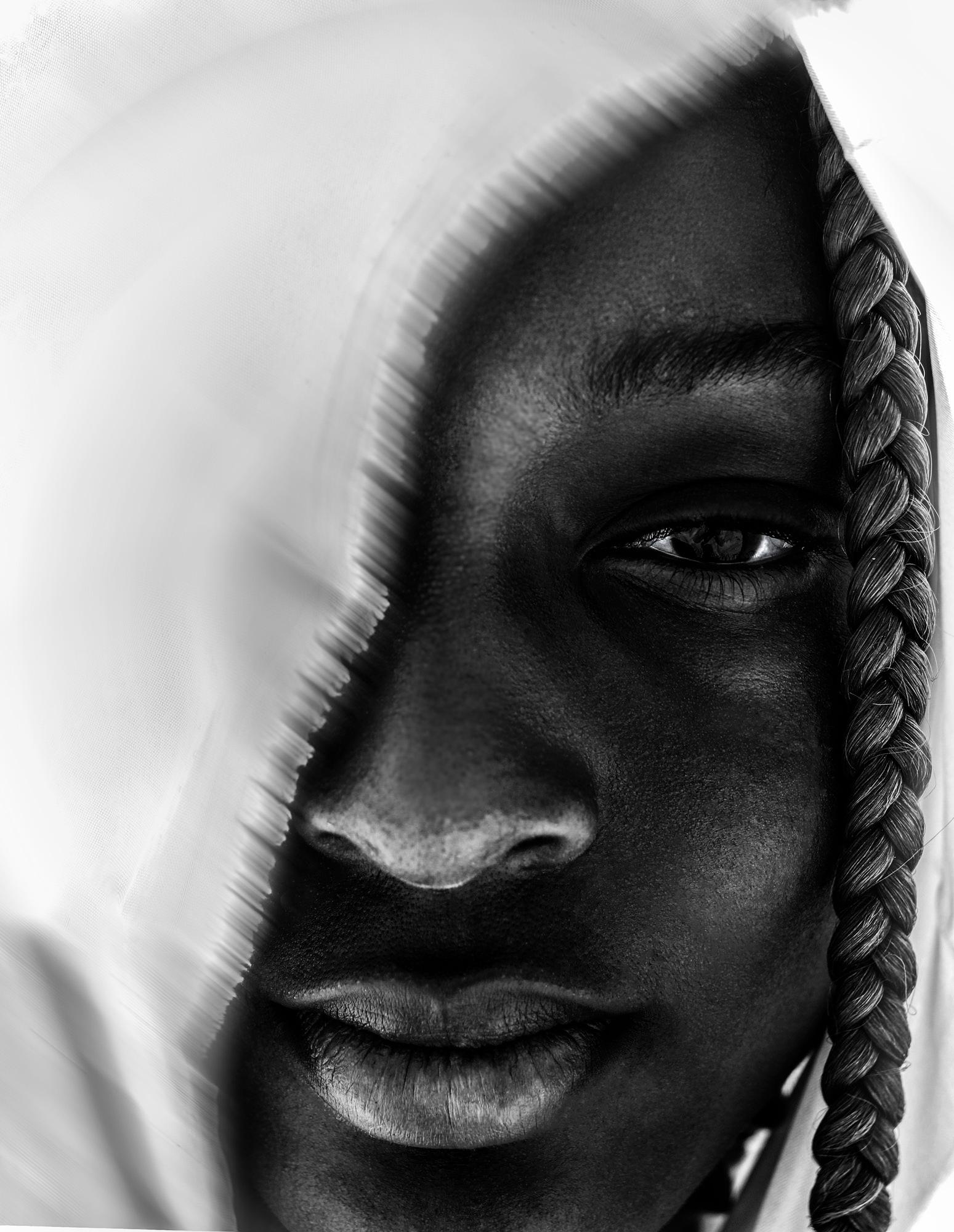 A close-up of a person's face, with a white cloth covering their head and a braid of hair visible on the side of their face.