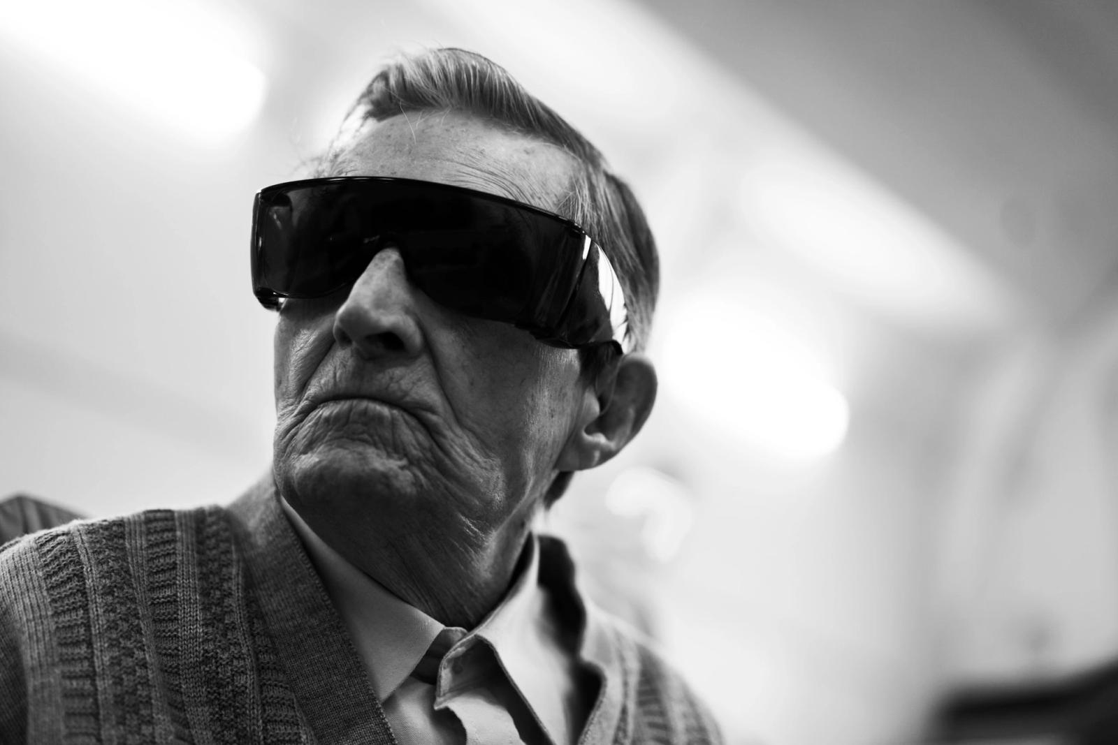 An older man with short hair wears a dark sweater over a collared shirt and dark sunglasses.