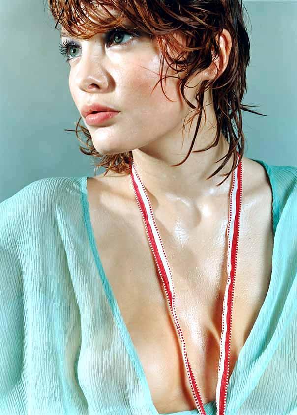 A woman with short, curly brown hair and a light blue blouse with a deep V-neck, looking directly at the camera with a subtle, introspective expression.
