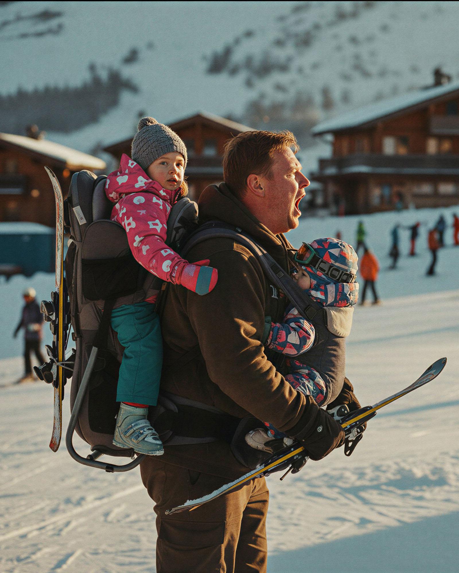 A man carrying two children on his back, both wearing winter clothing, in a snowy mountainous landscape with ski resorts in the background.