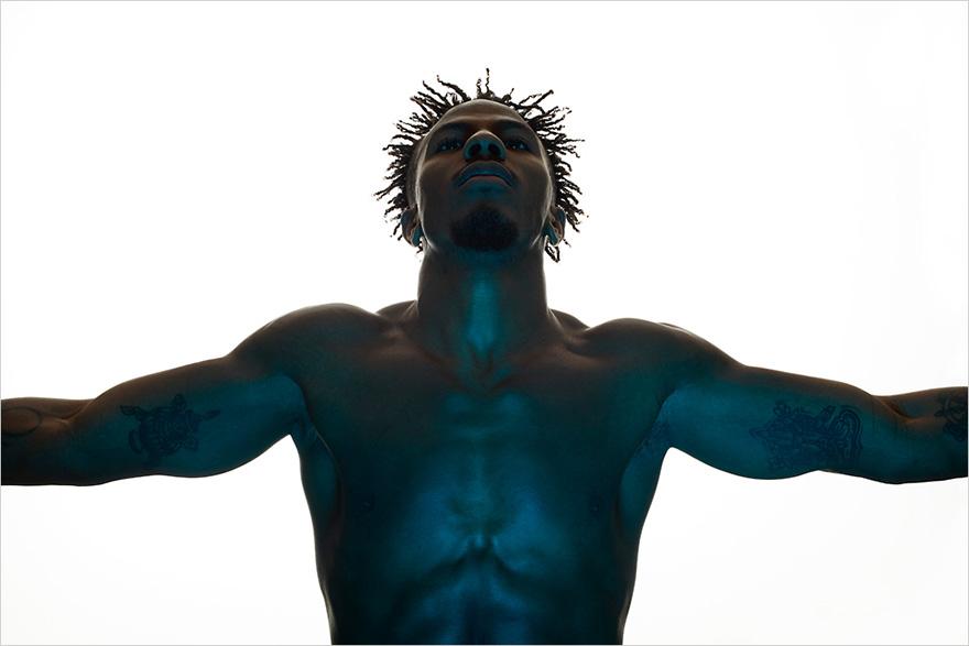 A shirtless, muscular black man with dreadlocks and tattoos, standing with arms outstretched and looking up at the sky.
