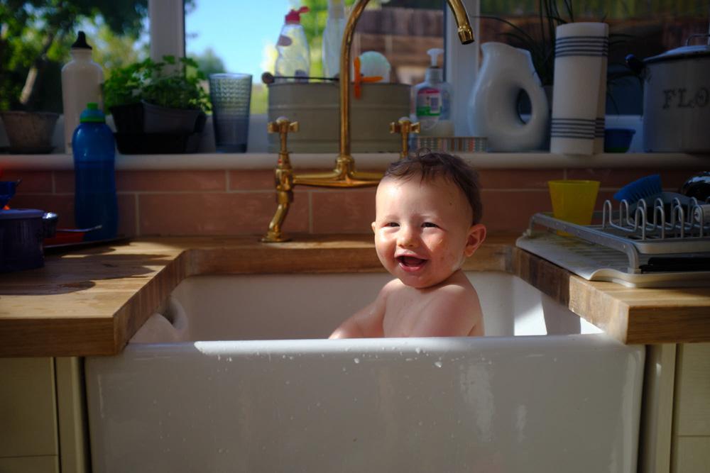 A baby sits in a white sink, smiling at the camera with a happy, relaxed expression. The sink is in a well-lit, sunny kitchen with a gold faucet and various kitchen items in the.
