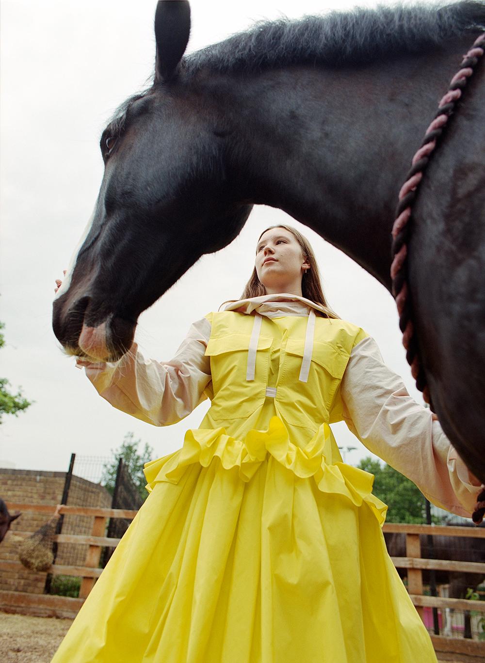 A woman in a yellow dress is standing in a stable, surrounded by horses, holding a horse's head.