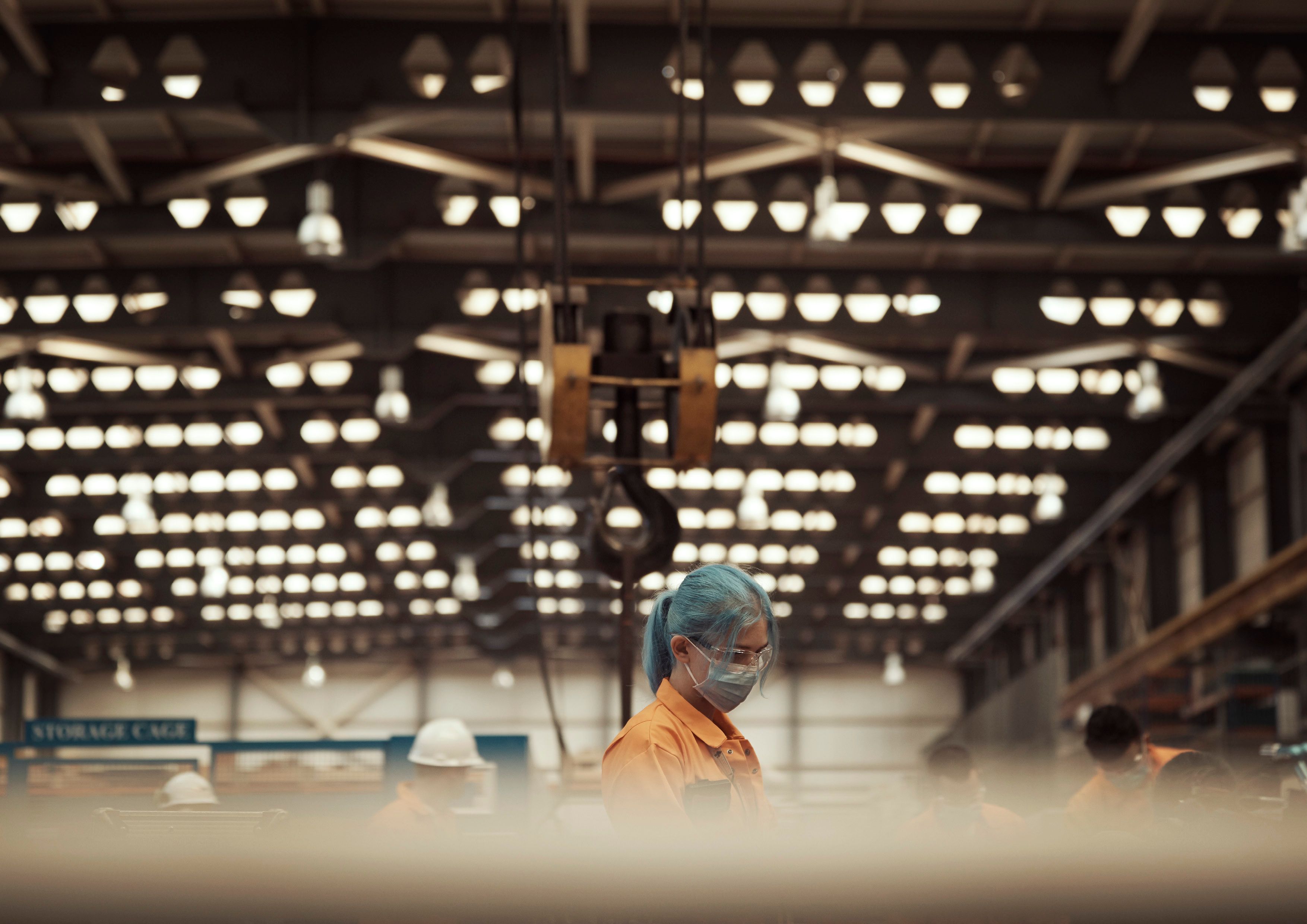 A woman with blue hair and a mask among workers in an industrial warehouse with overhead lights.