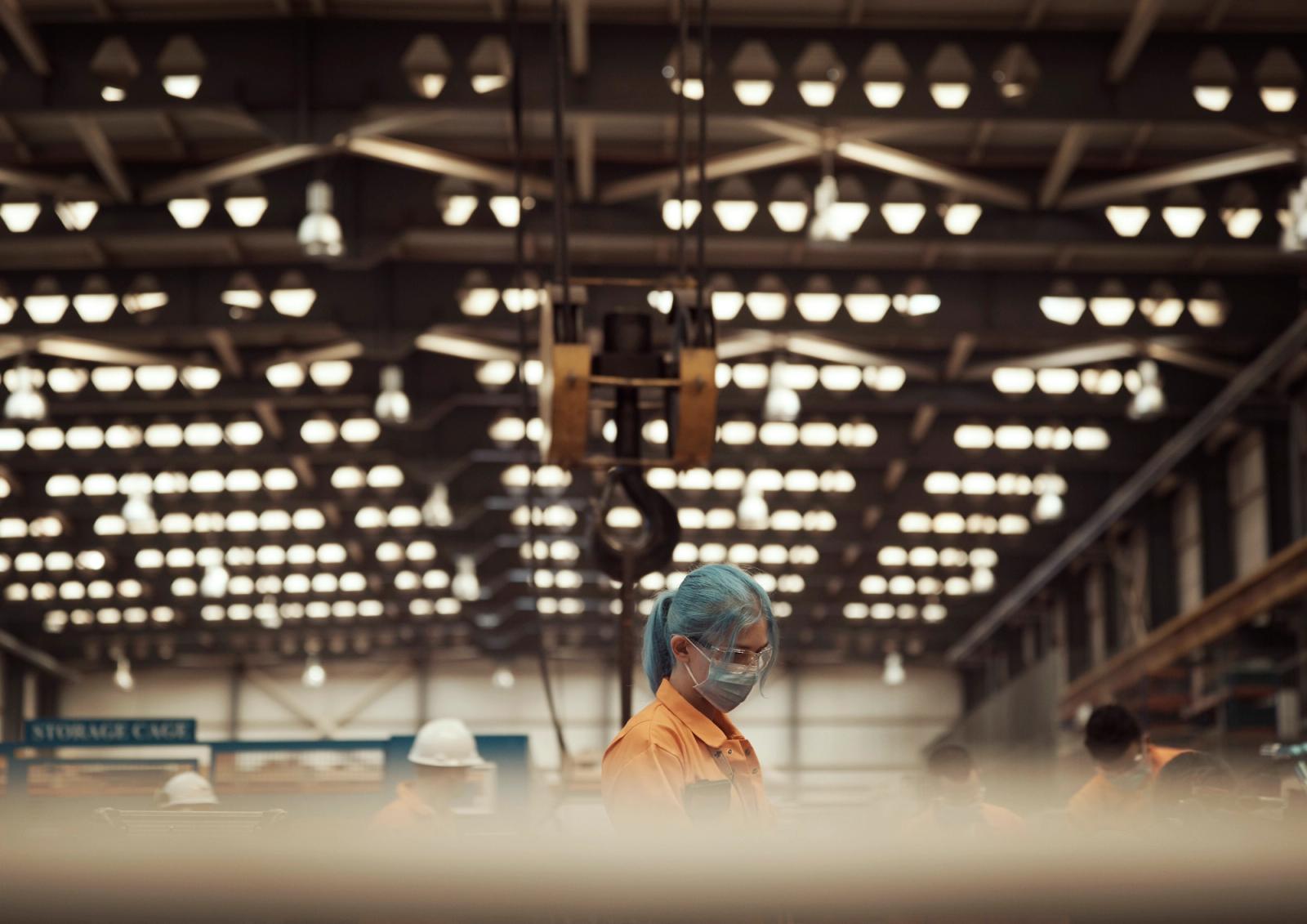 A woman with blue hair and a mask among workers in an industrial warehouse with overhead lights.