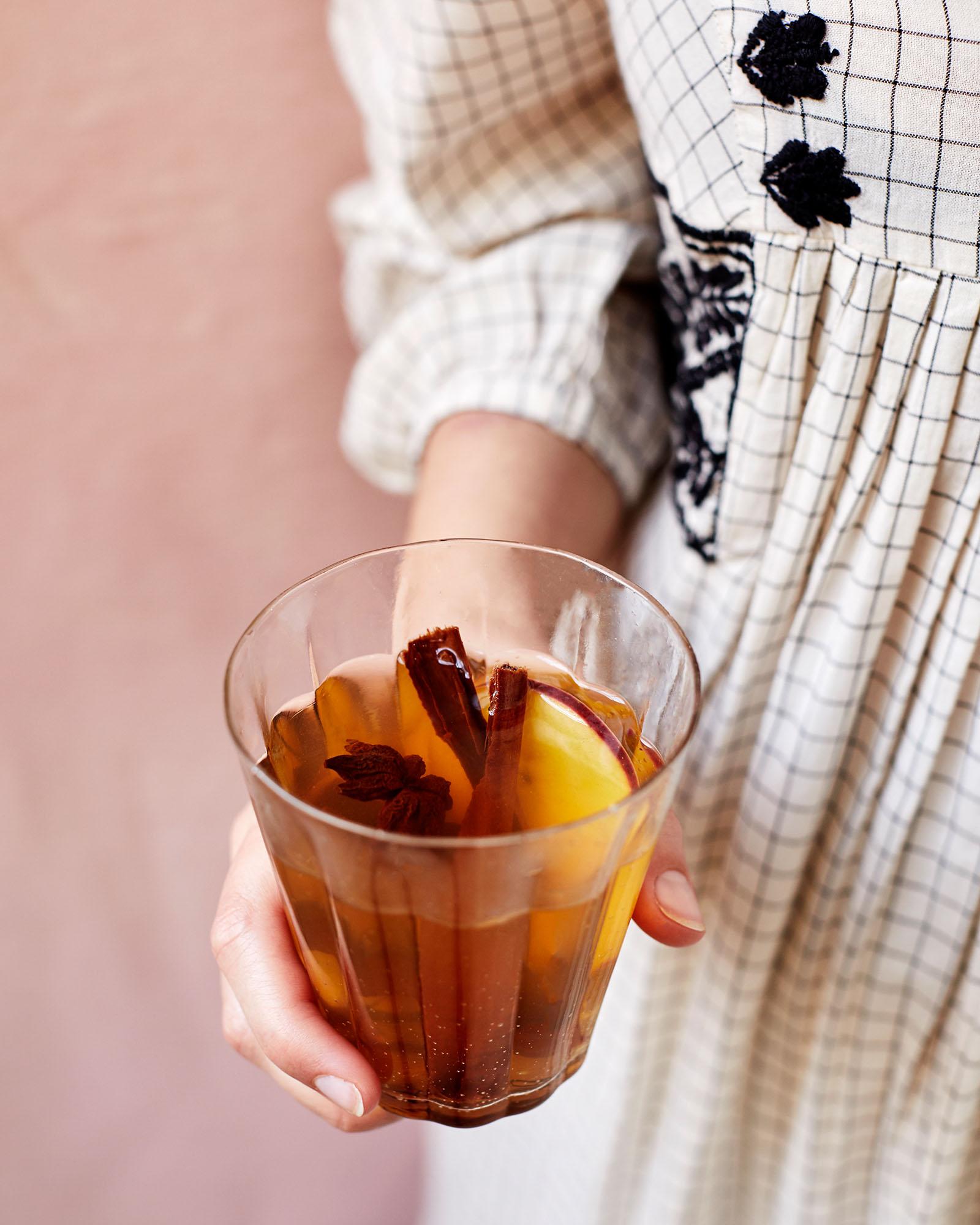 A woman is holding a glass of mulled wine, with a cinnamon stick and a few apple slices in it, in front of a warm, autumnal background.