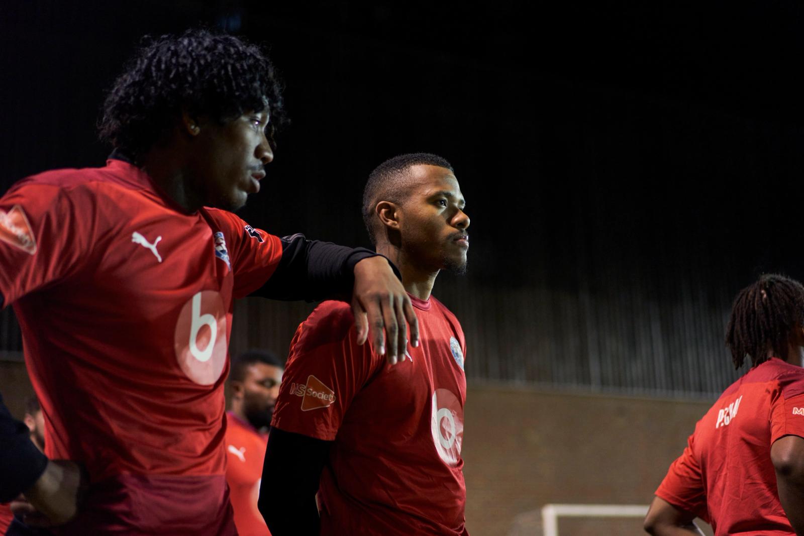 A candid shot of two football players in matching red jerseys standing together under night lights. One player rests an arm on the other's shoulder as they both look intently into the distance. Photograph by Nick Dolding.