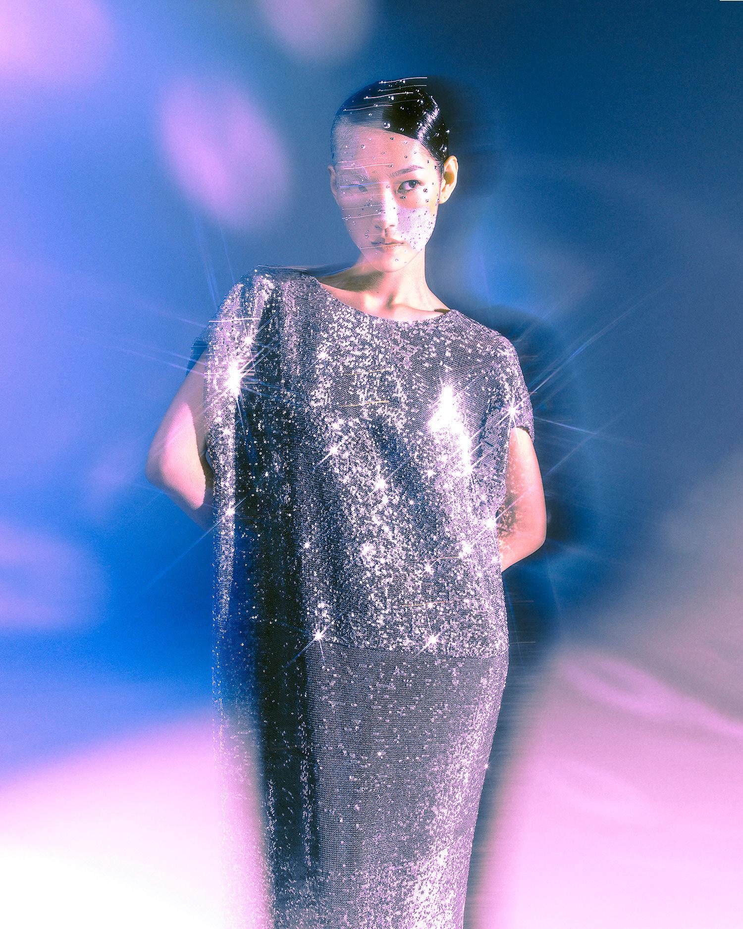 A woman wearing a long, dark, sequined evening gown with a high neckline and short sleeves, posing in a studio setting with a dark background.