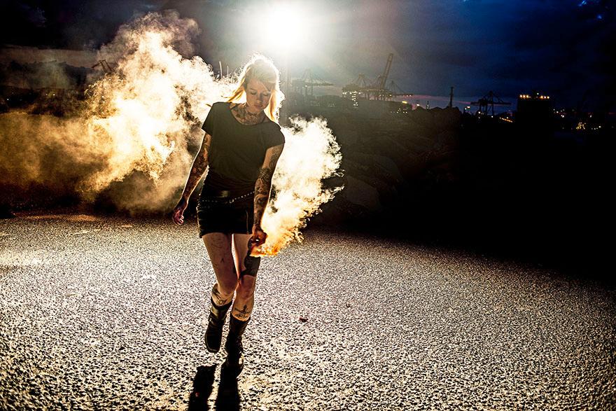 A woman in a black t-shirt and shorts, with tattoos, walking in a dark industrial area with a lit flare in her hand, surrounded by smoke.