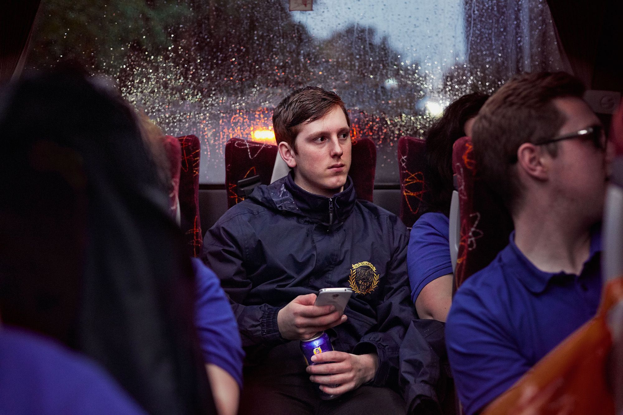 A man in a dark blue jacket with a yellow logo on the chest is sitting on a bus, looking out the window at a rainy city street.