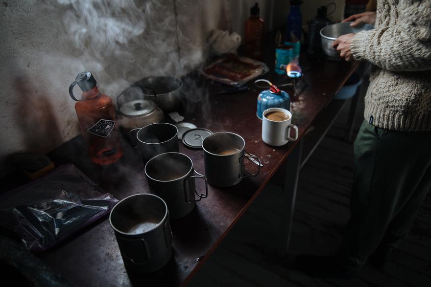 A person in a sweater and trousers is standing in a small, dimly lit room with a table and several cups of steaming hot drinks.