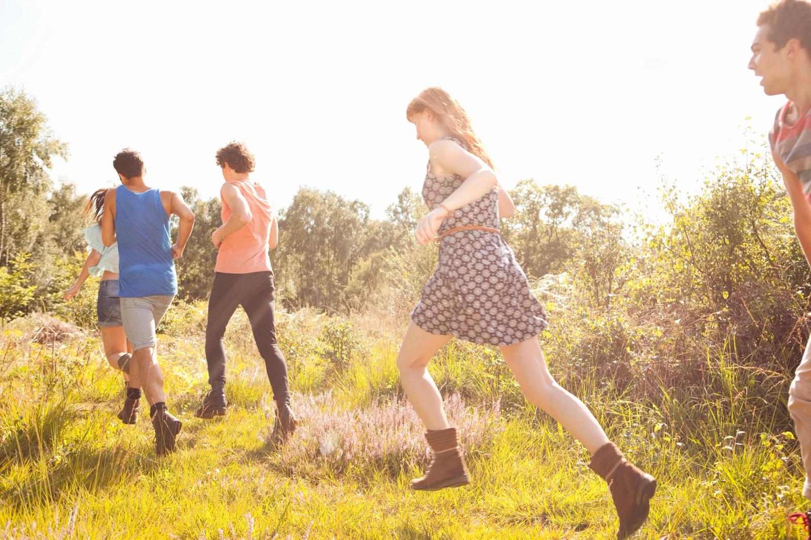 A group of people running through a field of tall grass on a sunny day, wearing casual clothing and boots.