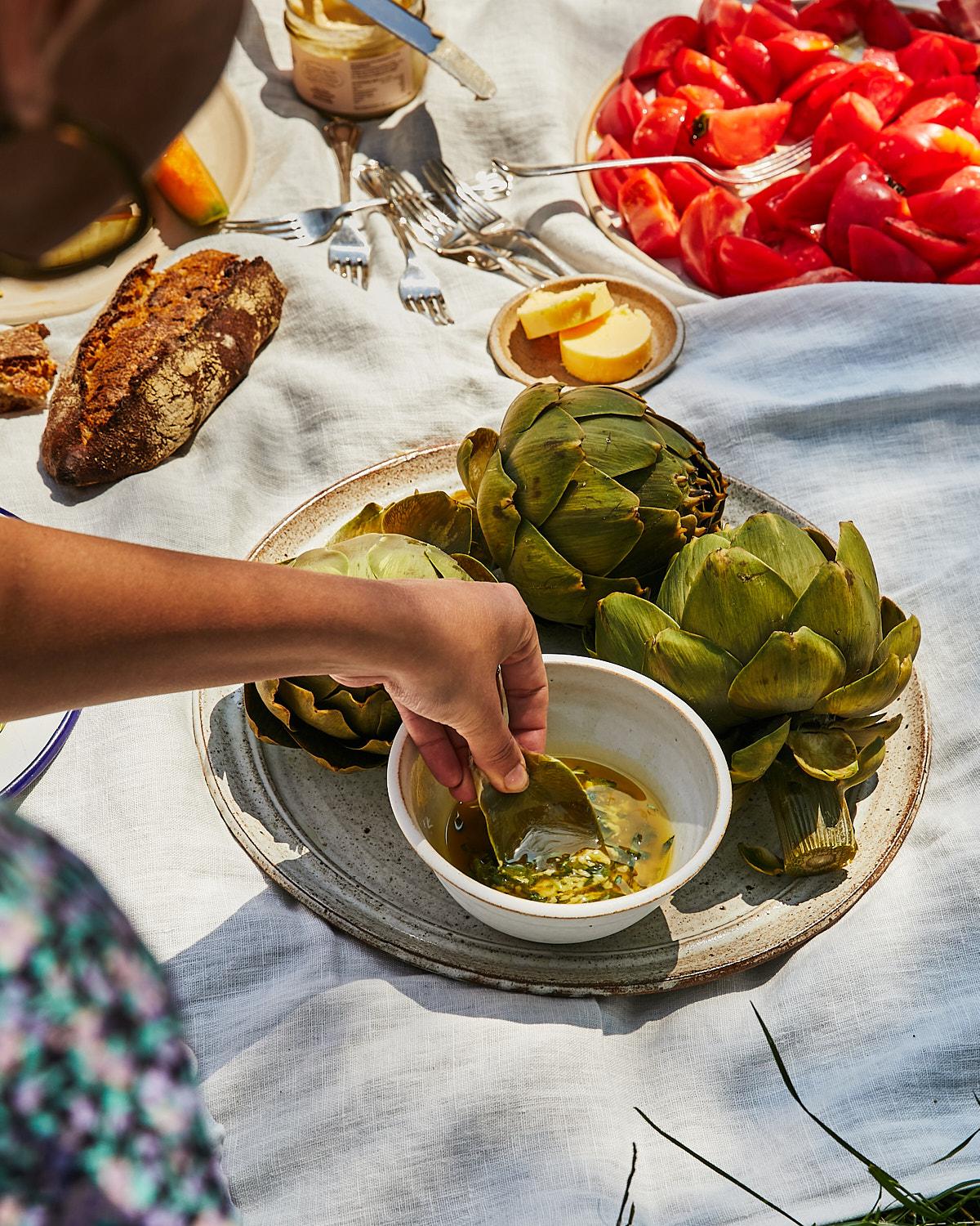 A person's hand is reaching into a bowl of artichokes, with a few artichokes on a plate in the background.