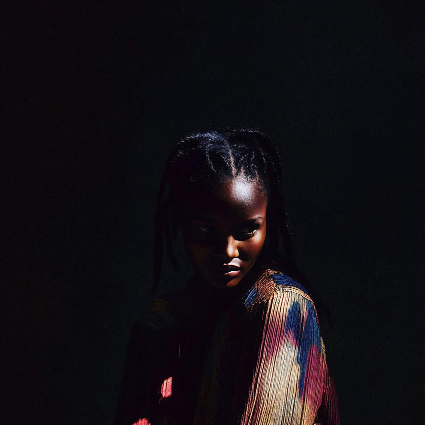 A young girl with braided hair and a colorful, striped shirt, standing in a dark room with a subtle, warm light on her face.