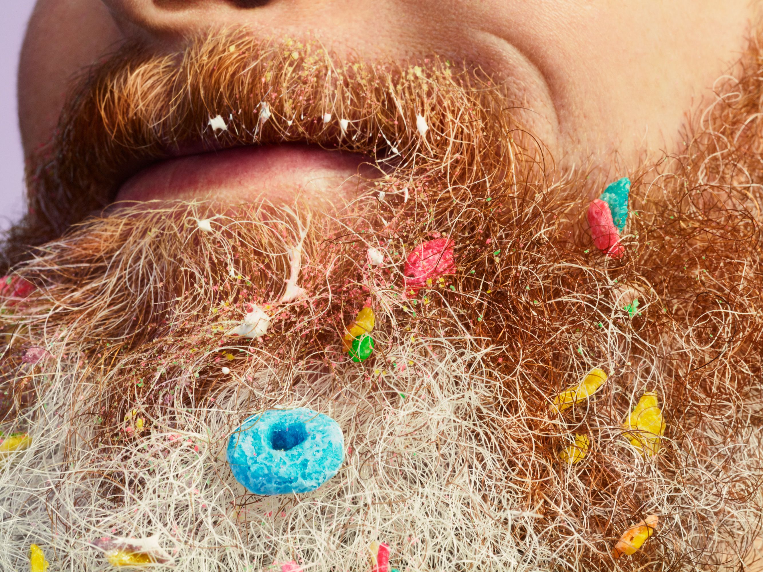 A close-up of a man's face with a thick, messy, and colorful beard, with various food items and other objects stuck in it.