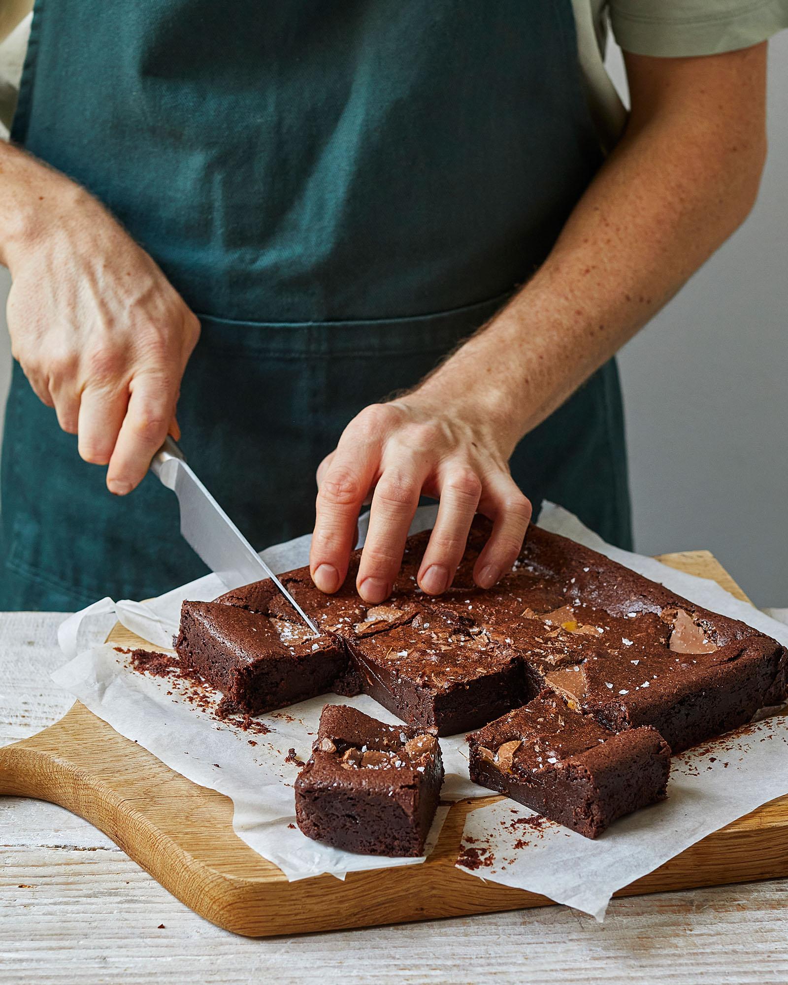 A person in a green apron is cutting a brownie on a wooden cutting board.