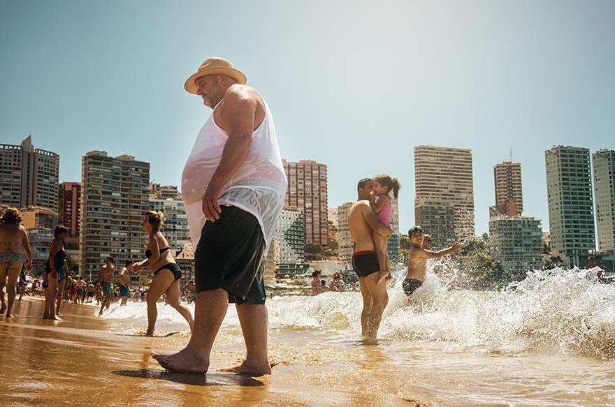 A man in a white tank top and black shorts walking on a beach with a city skyline in the background.