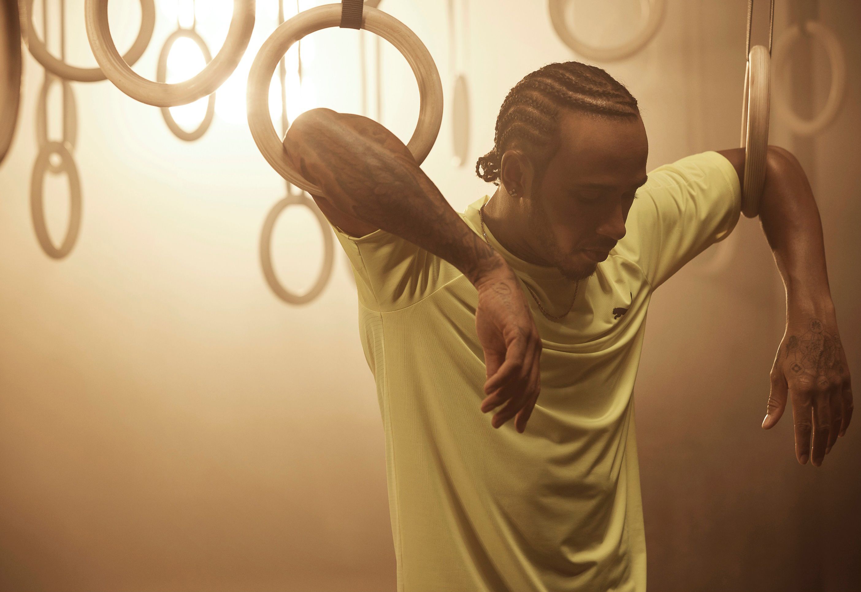 A man with tattoos and braided hair is posing in a gym, wearing a yellow t-shirt and holding onto gymnastics rings.