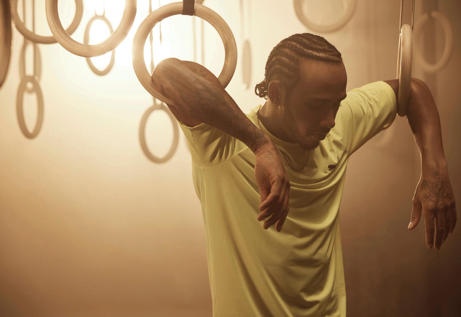 A man with tattoos and braided hair is posing in a gym, wearing a yellow t-shirt and holding onto gymnastics rings.