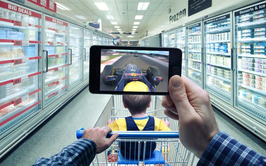 A person holding a smartphone, taking a photo of a Formula 1 car racing on a screen in a supermarket aisle, with a child in a shopping cart in the foreground.