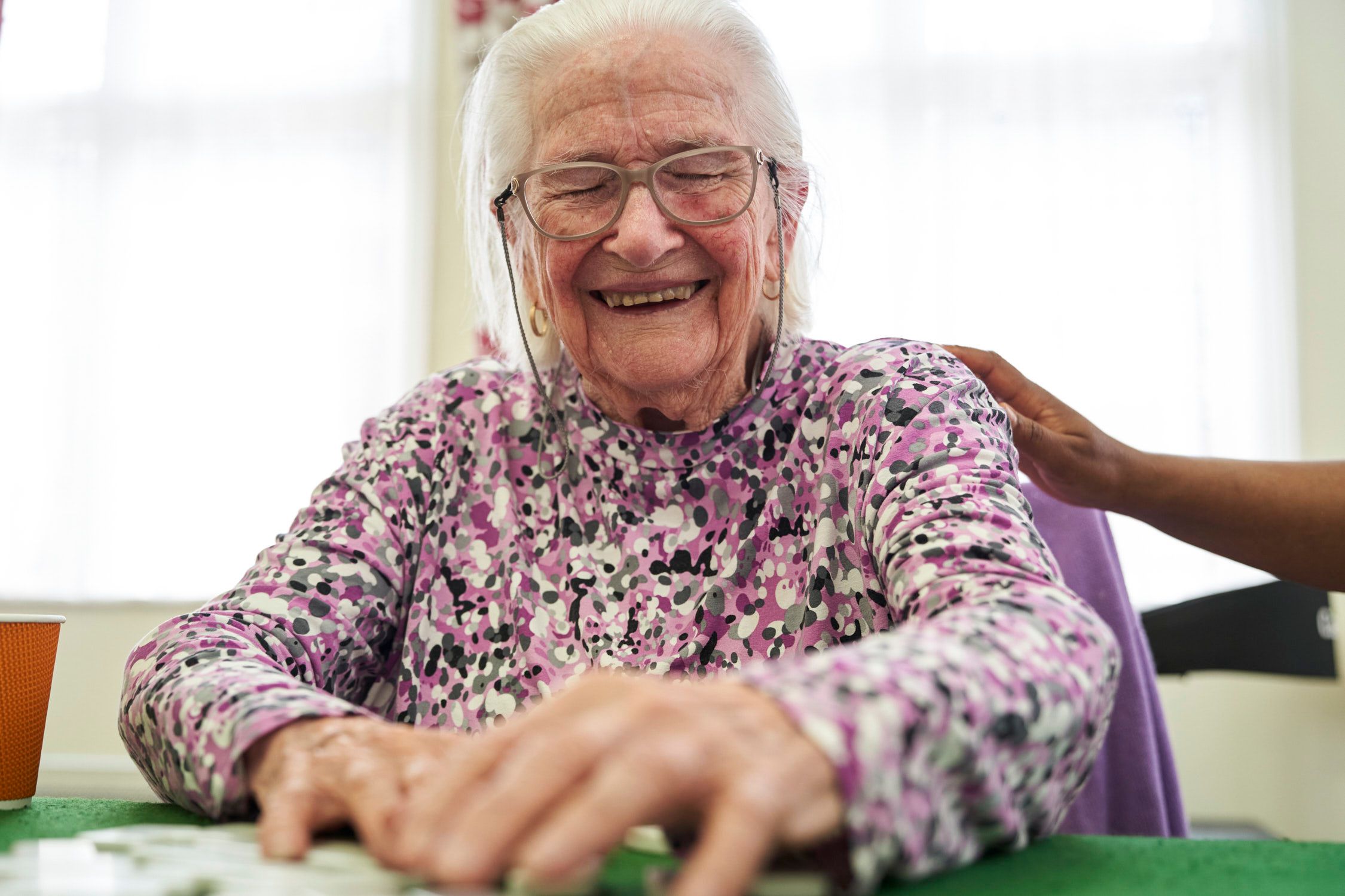 An elderly woman with white hair and a purple floral blouse is smiling and playing a game of cards.