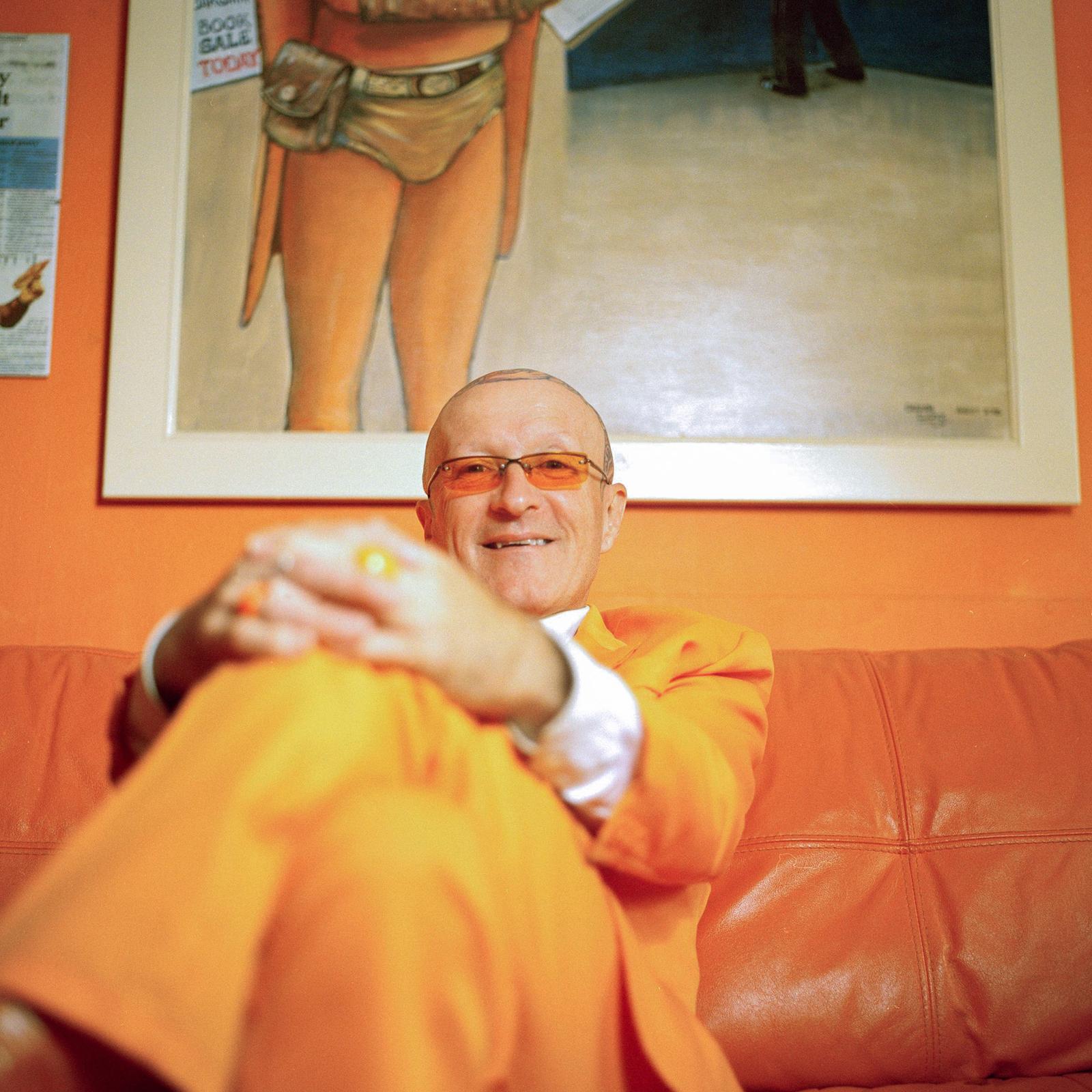A man sits on a couch wearing a bright orange suit, with a painting of a woman in a red dress visible behind him.