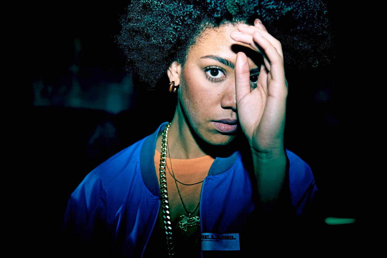 A woman with curly hair and a blue jacket, looking directly at the camera with a serious expression.